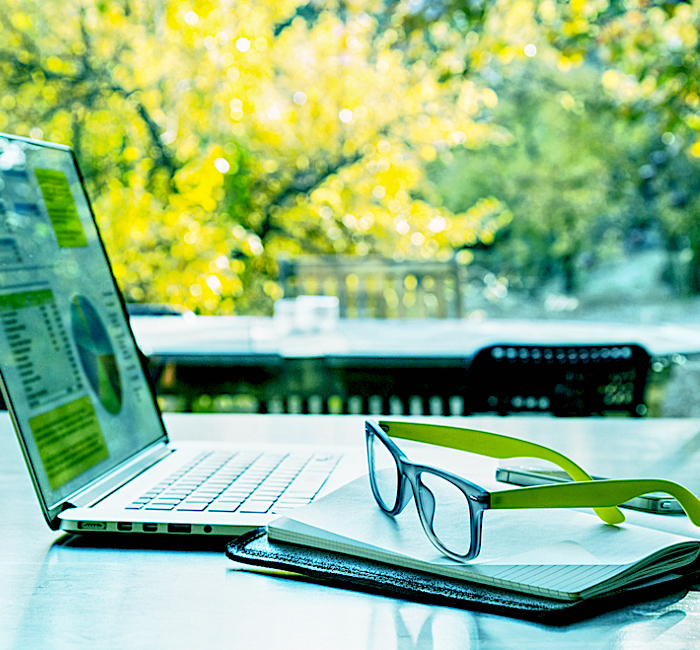 Laptop auf einem Tisch draußen in der Natur. Im Hintergrund sind Bäume mit grünem und gelbem Laub zu sehen.